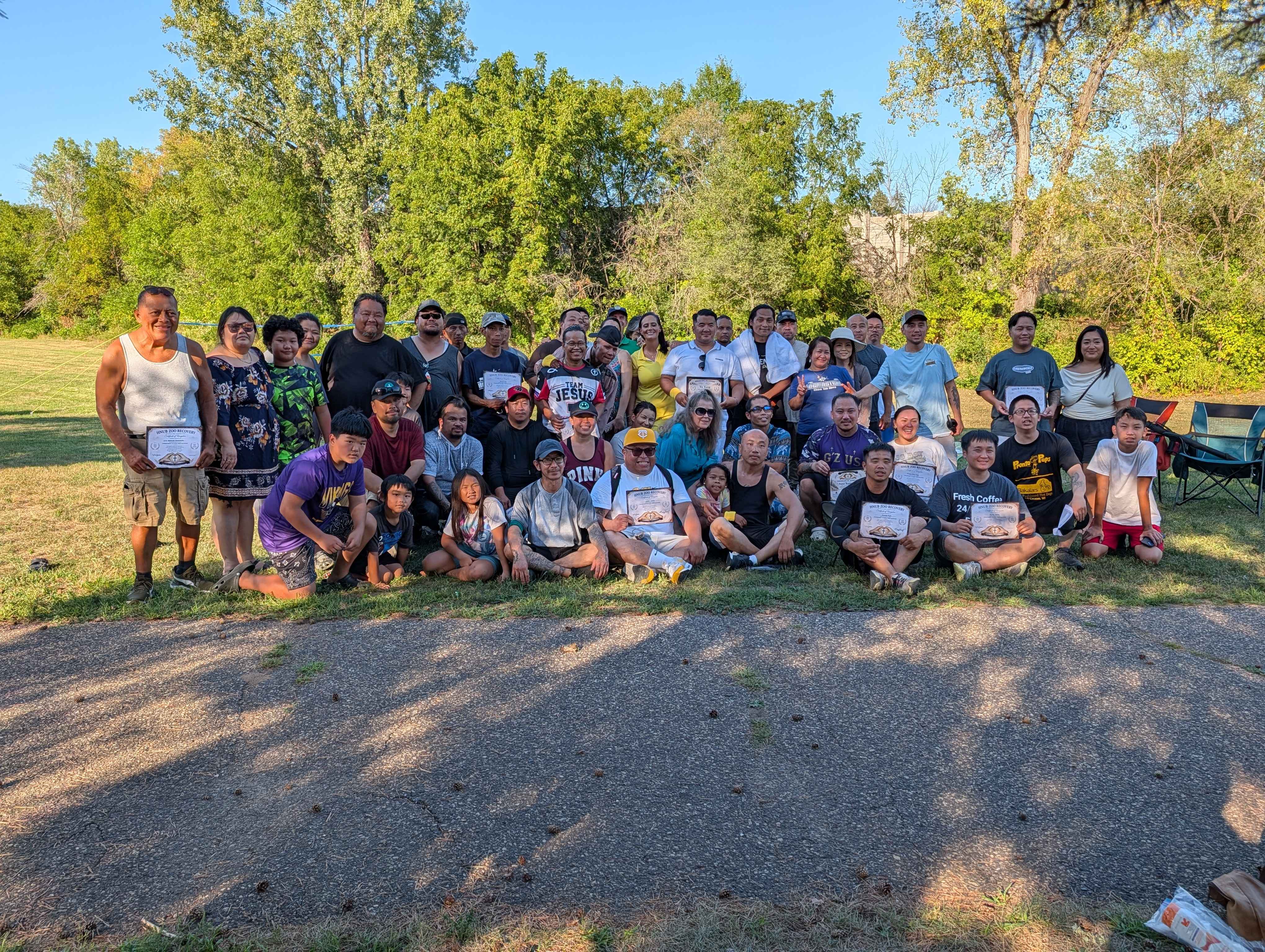 A large group of Housing Support Rides clients, volunteers, and staff gathered outdoors, several holding recovery milestone certificates